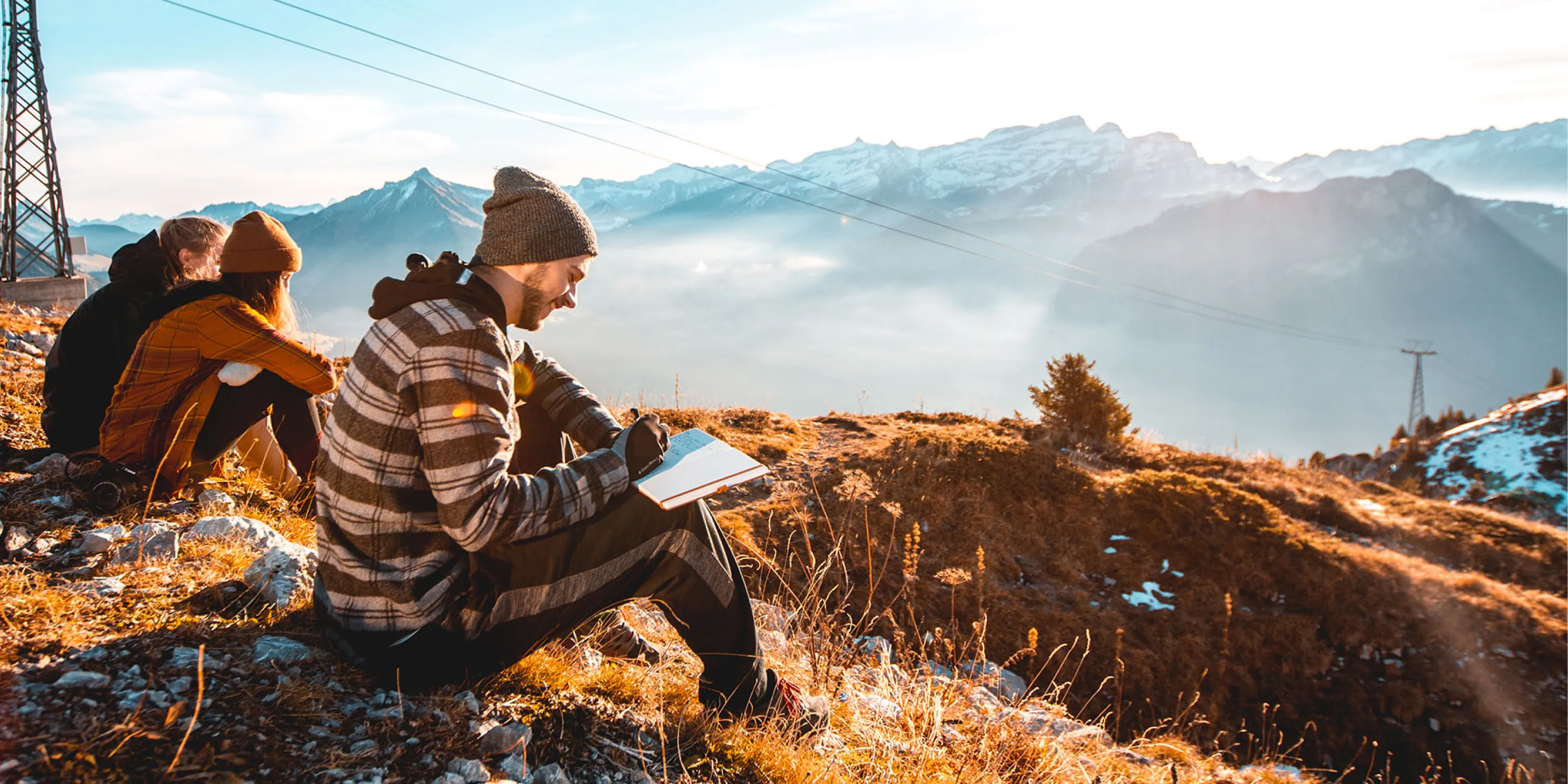 A couple of students sitting on a mountain overlooking the view with one man writing notes in a journal.