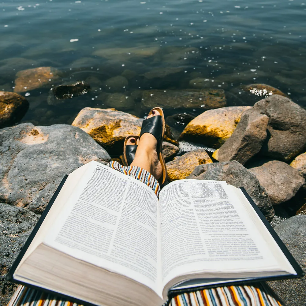 An image of a bible laid out on a student's lap overlooking rocks and water.
