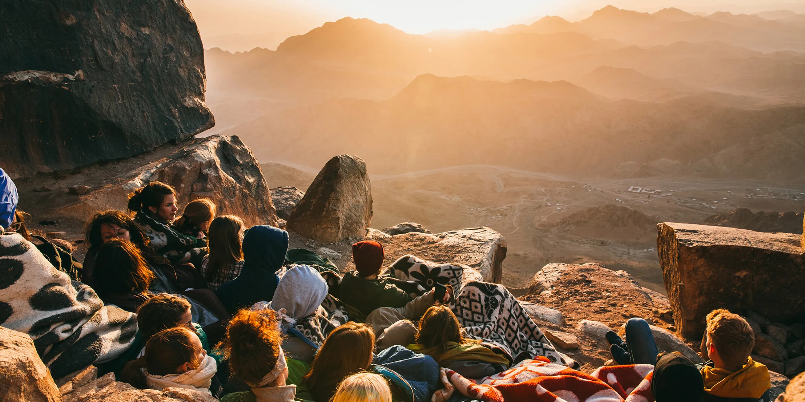 A group of male and female students lying on a mountain with blankets overlooking the sunrise.