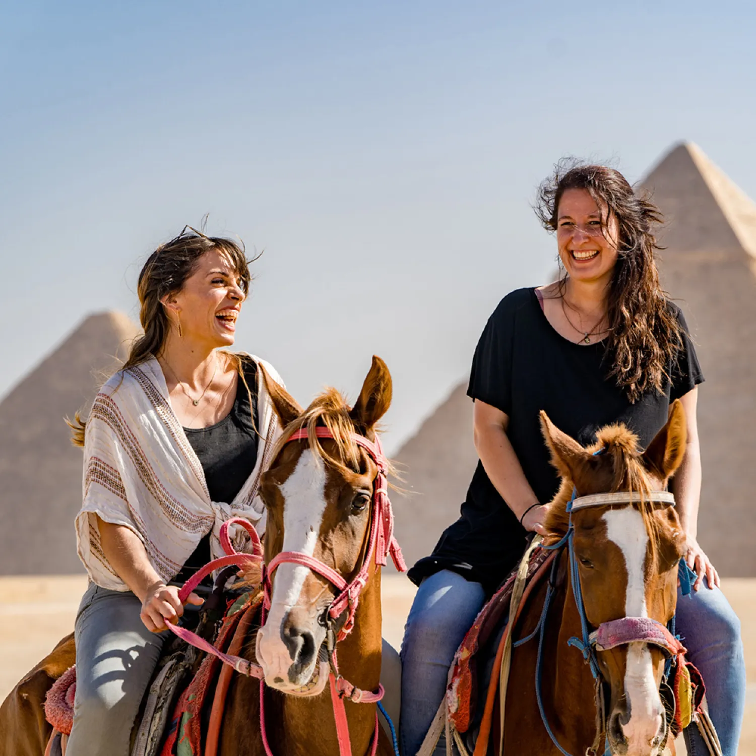 Two women smiling and sitting on horses with pyramids in the background.