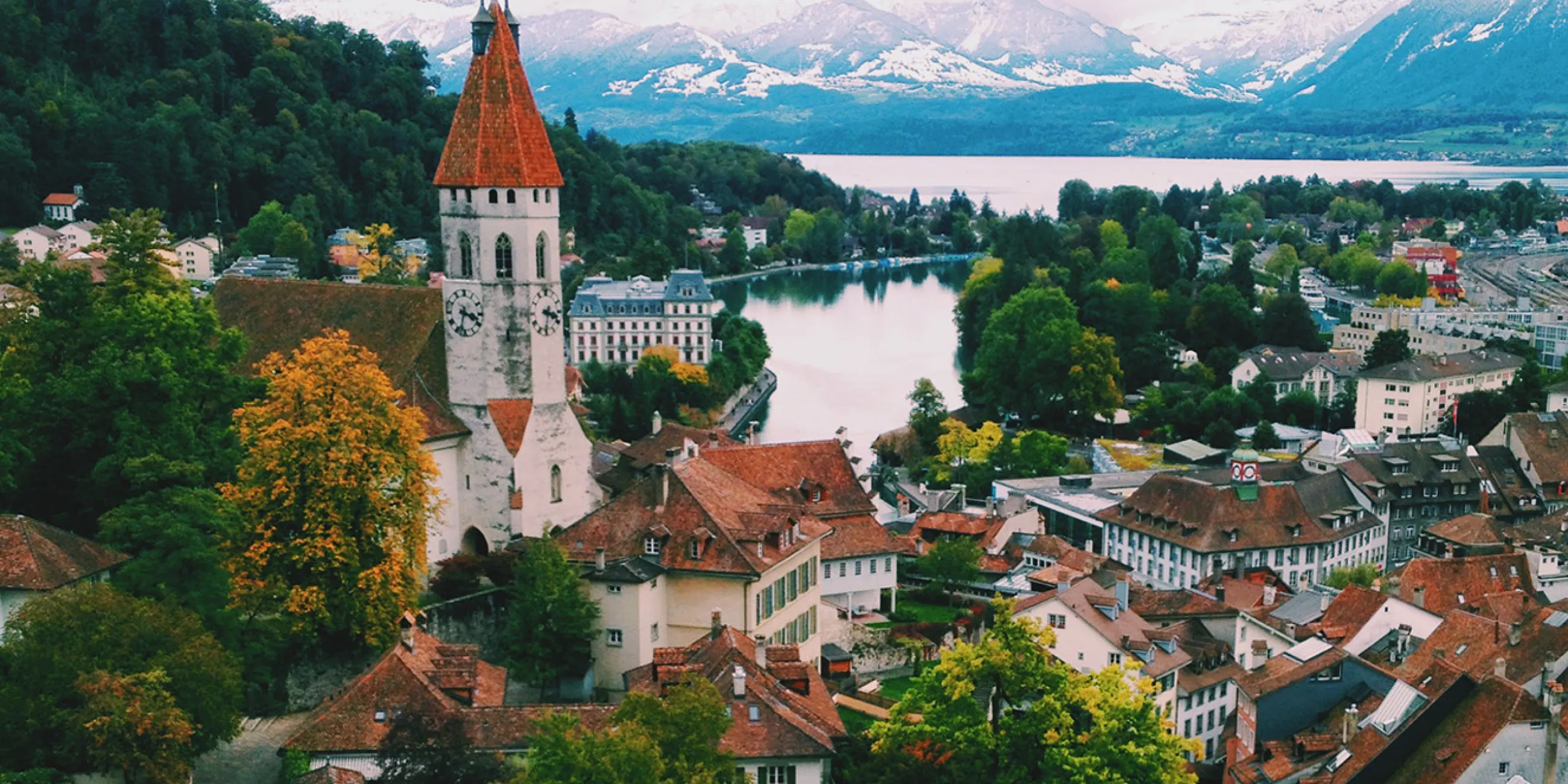 A landscape image of Switzerland containing architecture, a lake, greenery, and snow-capped mountains.