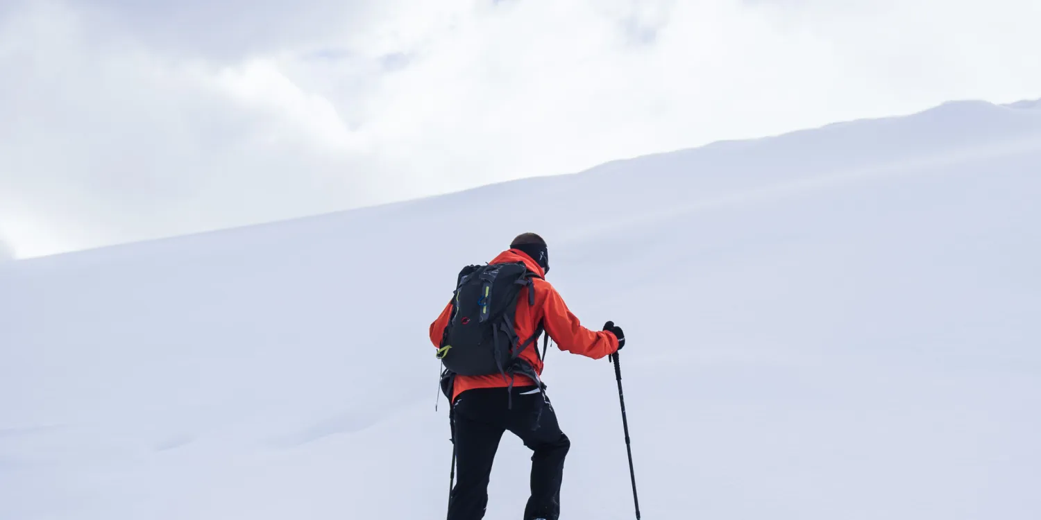 A man in a red jacket hiking up a hill of white snow.