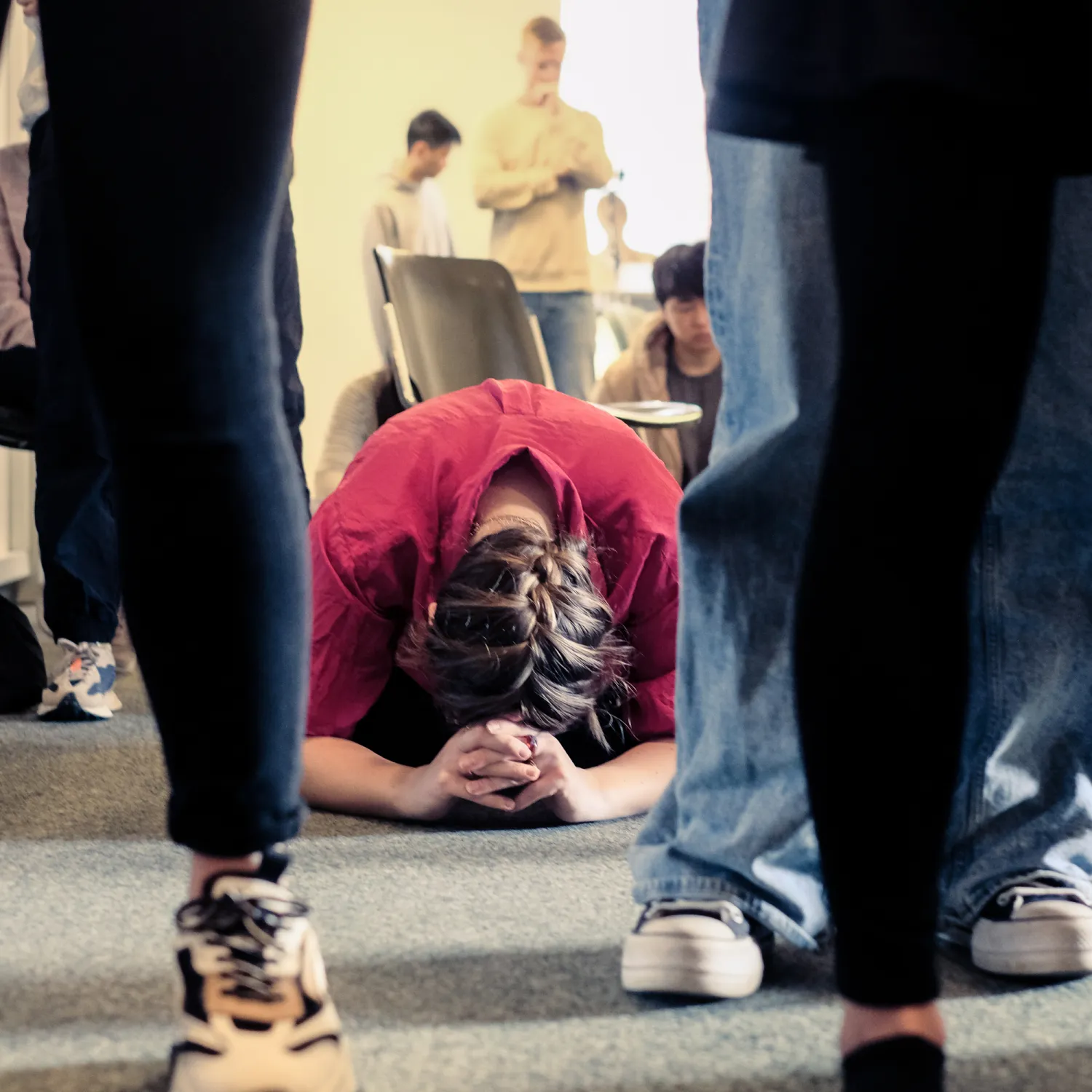 A student kneeling down on the ground among others in a classroom during worship school.