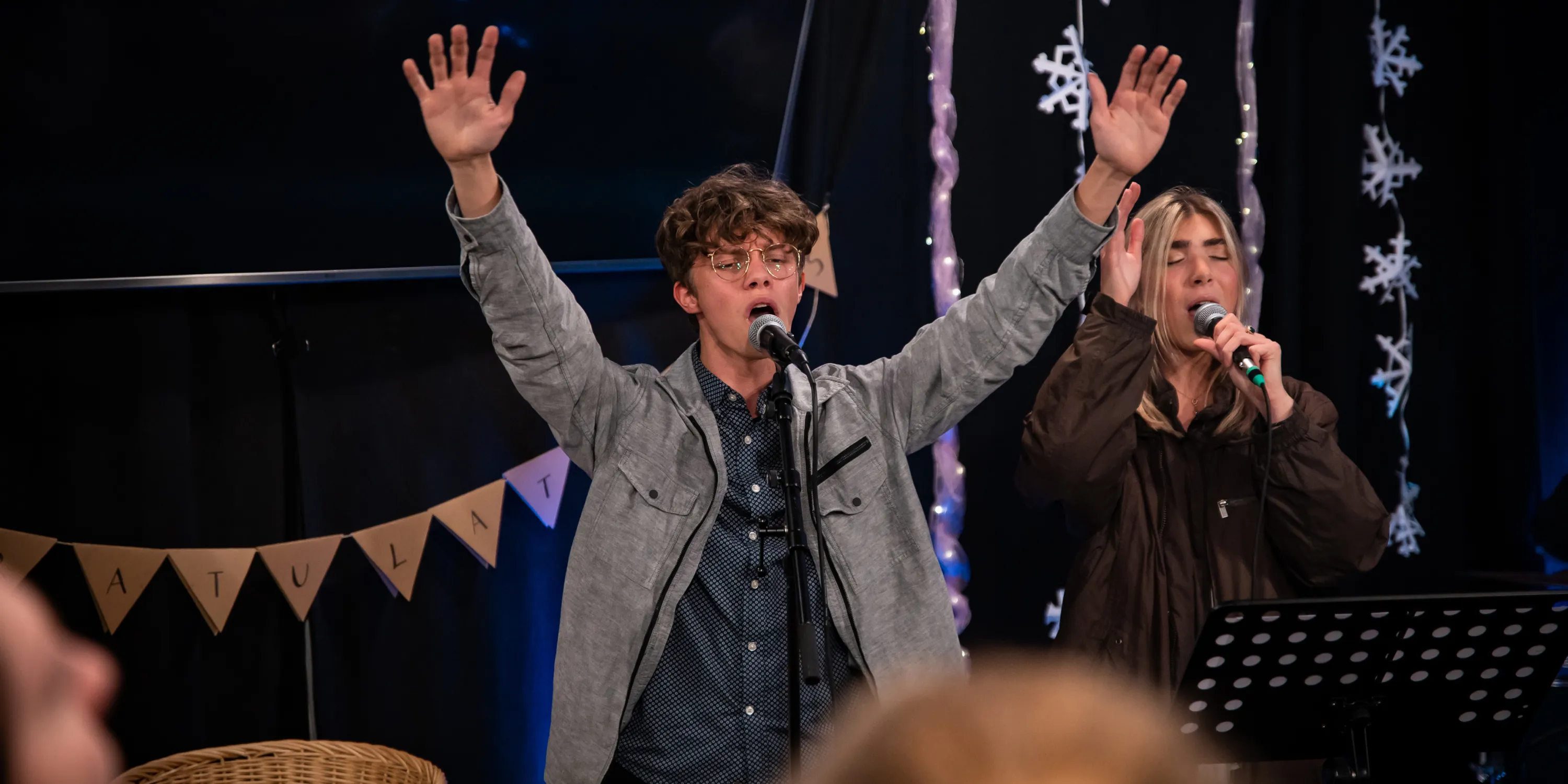Two students singing into microphones on a stage with their arms up in the air and eyes closed.
