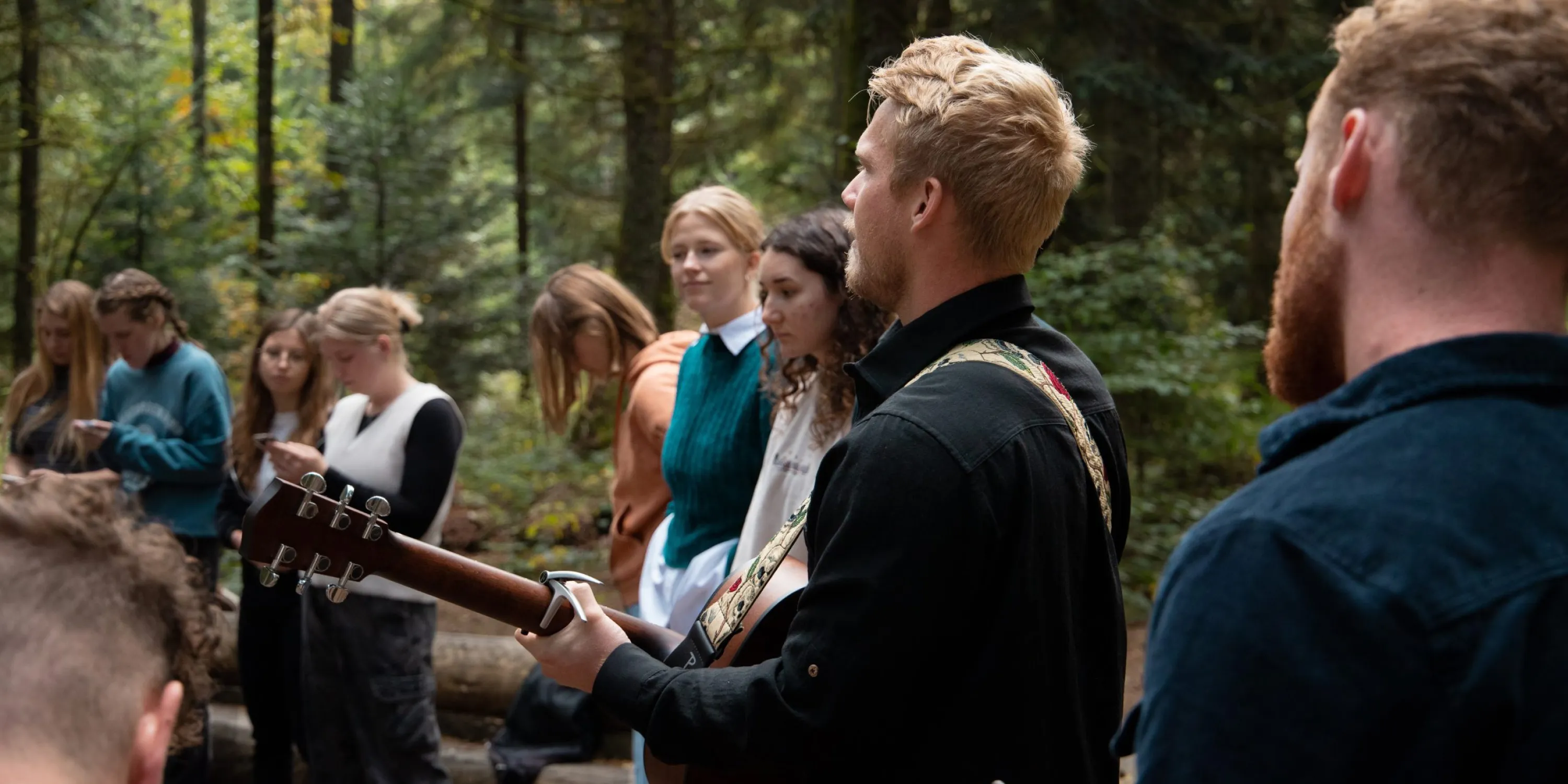 A group of students singing and playing guitar outside among the trees.