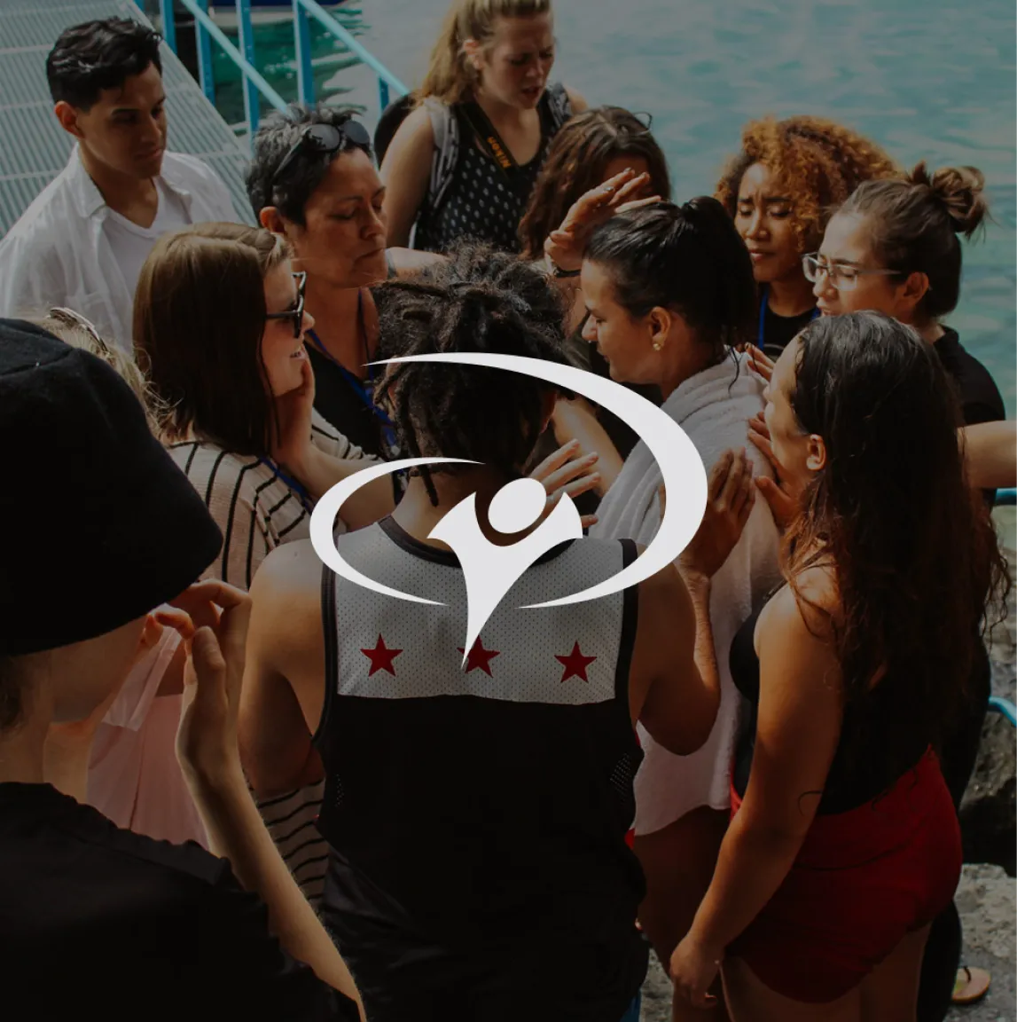 The YWAM logo overlaying an image of a crowd of gathered students praying with their hands placed on each others' backs.