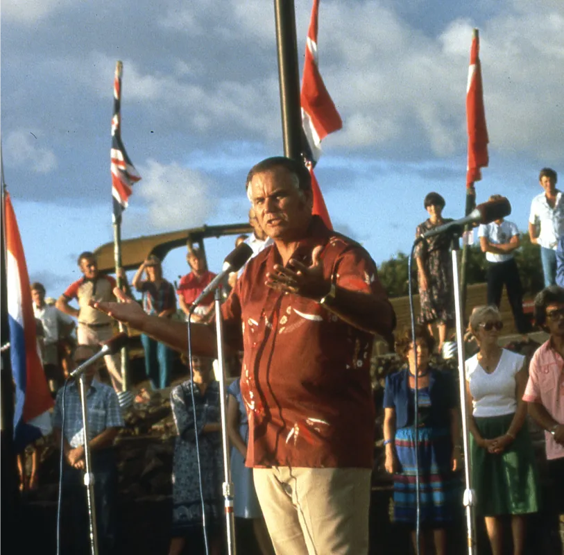 A man speaking in a microphone in front of a crowd of people with flags blowing in the background.
