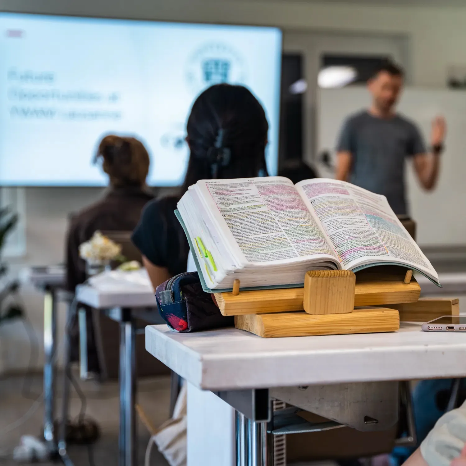 A bible laid out on a desk in a classroom with a teacher giving a lesson in the background during DTS at YWAM Lausanne.