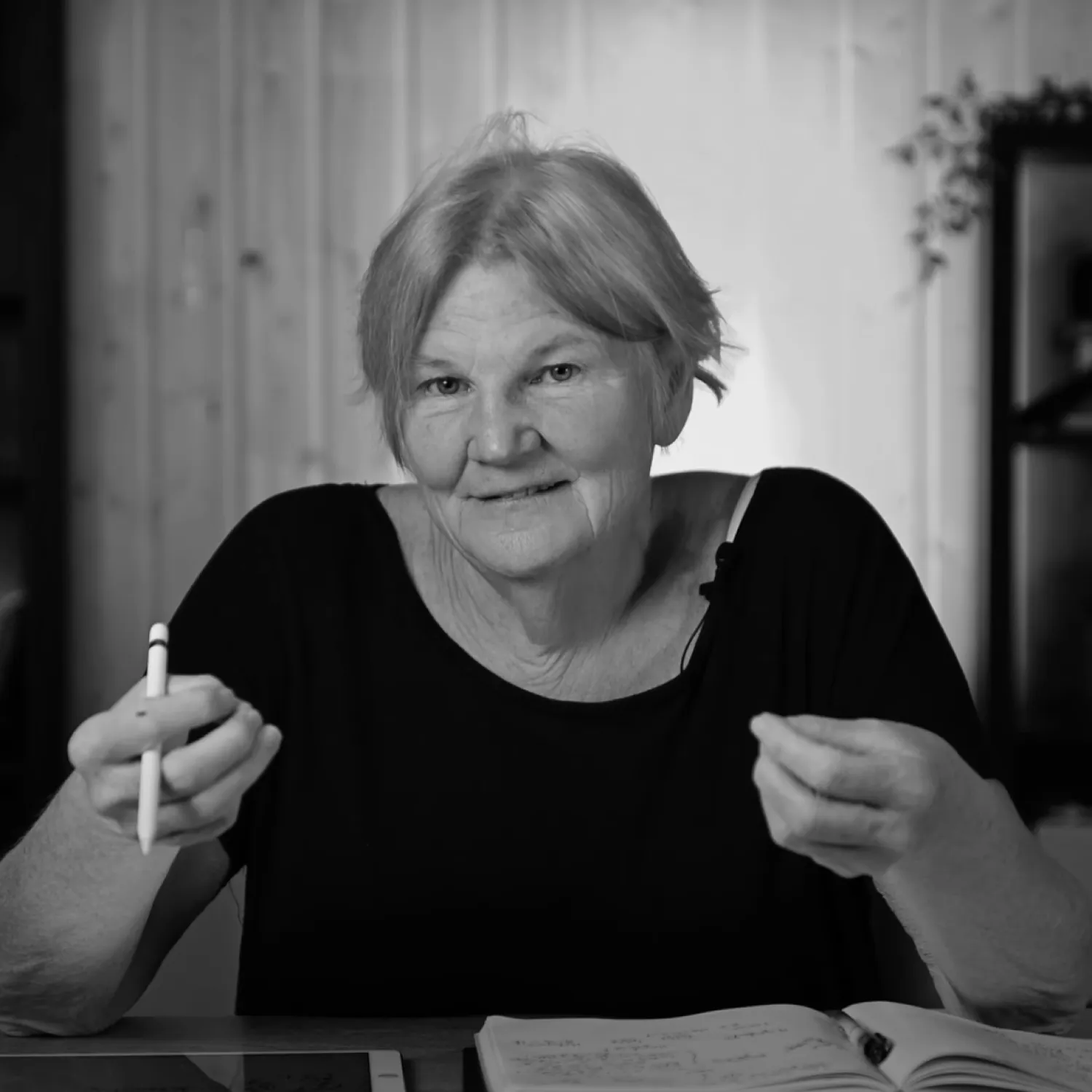 An older woman sitting at her desk talking while holding a pen.