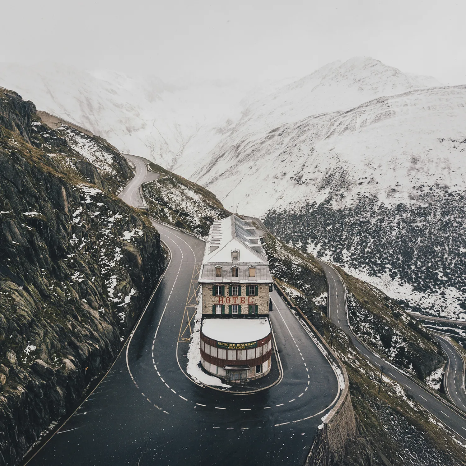 An overhead image of a building in the middle of a bended road among the snow-capped mountains.