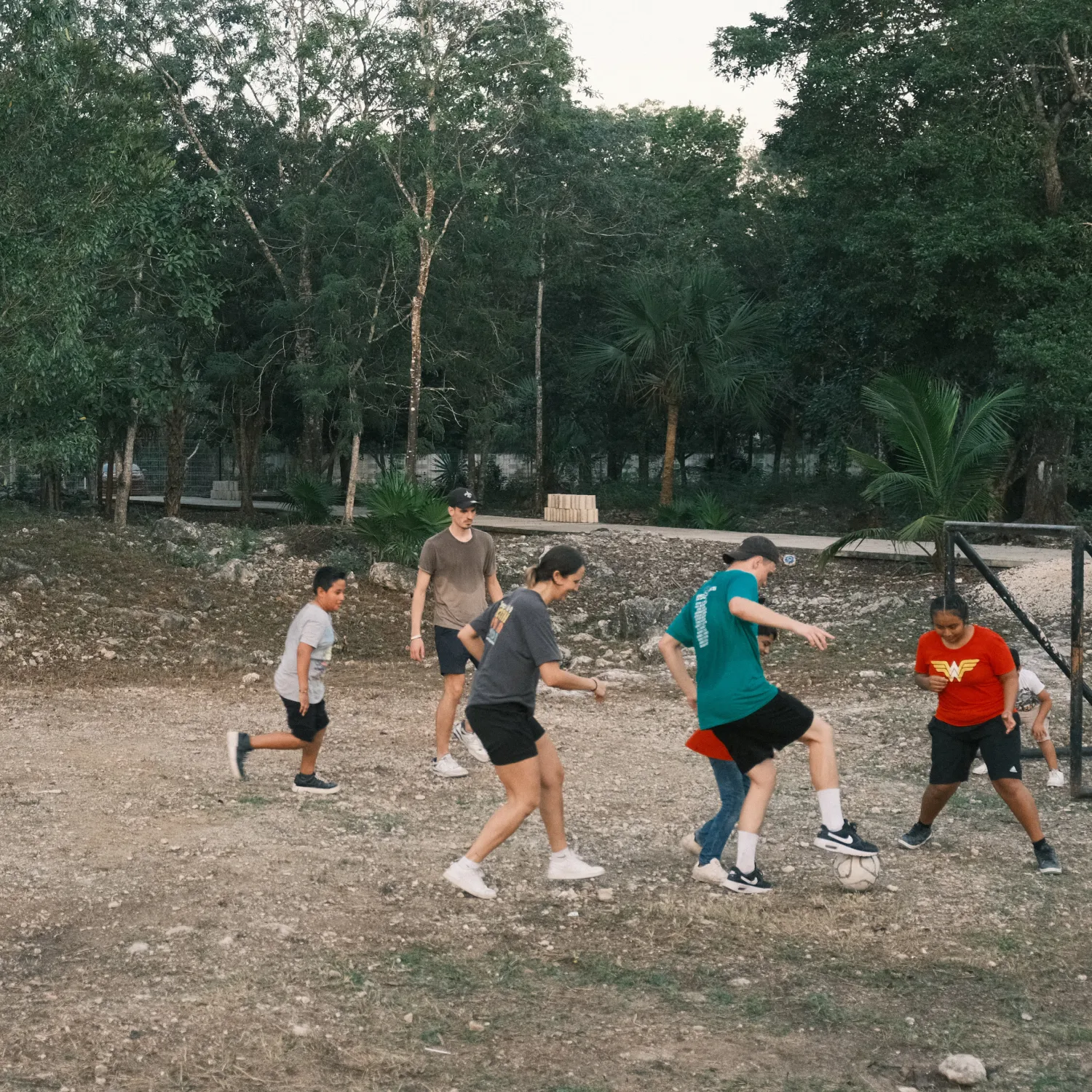 A group of children playing football on an outside sandy field with trees in the background.