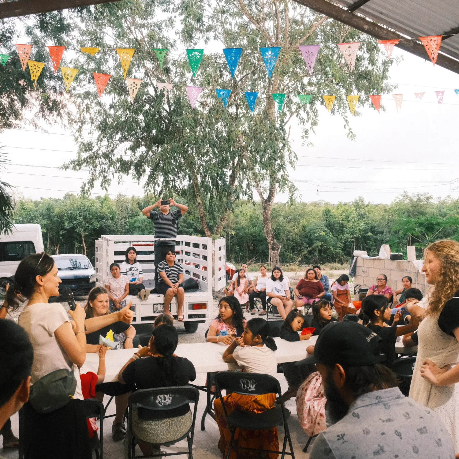 A group of children and adults sitting on chairs around a sheltered table outside among trees under strings of colourful flags.