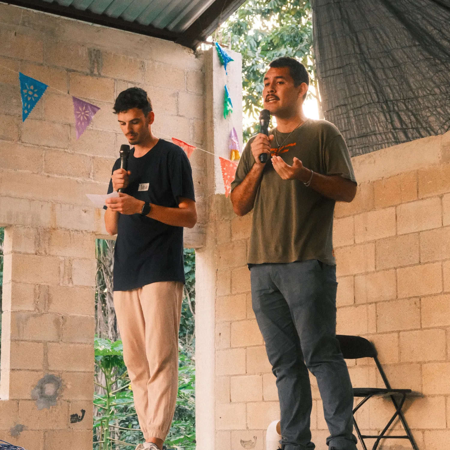 Two men standing on a stage and talking into microphones in a brick hall.