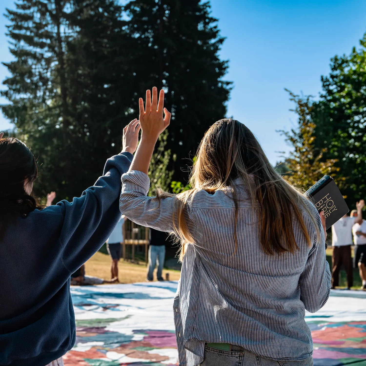 An image of a student from behind with one hand holding the bible and the other hand up in the air.