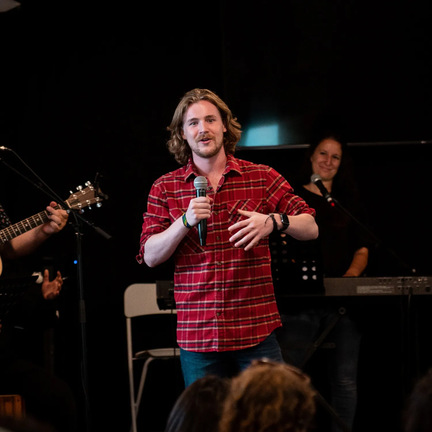 A man standing on a stage amongst musicians and speaking into a microphone in front of a group of people.