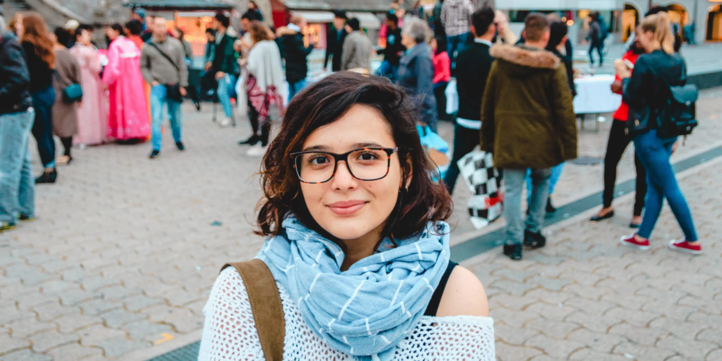 A student with glasses and a scarf smiling at the camera with a crowd of public people in the citybackground.