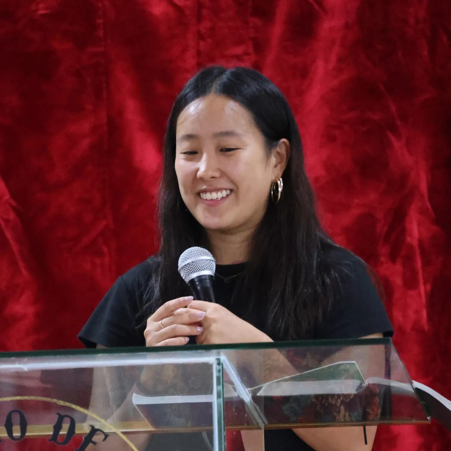 A student smiling and holding a microphone in front of a podium with a red velvet backdrop.