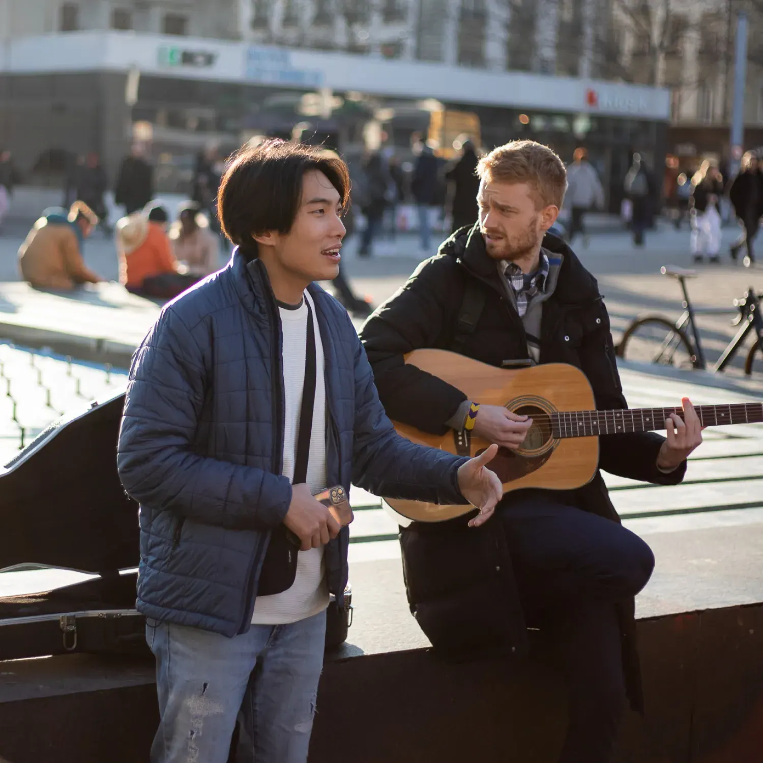 A man playing the guitar next to a man speaking with his arms out.