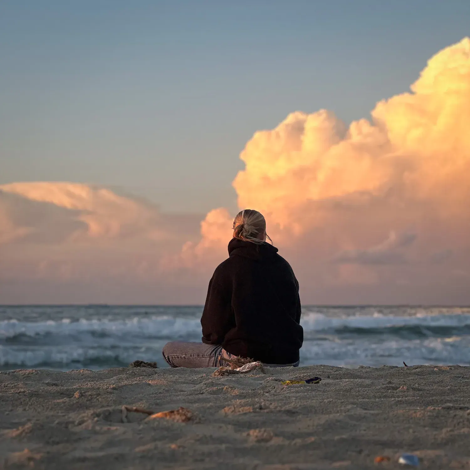 Person sitting on sandy beach facing the ocean under a sky with large orange clouds at sunset.