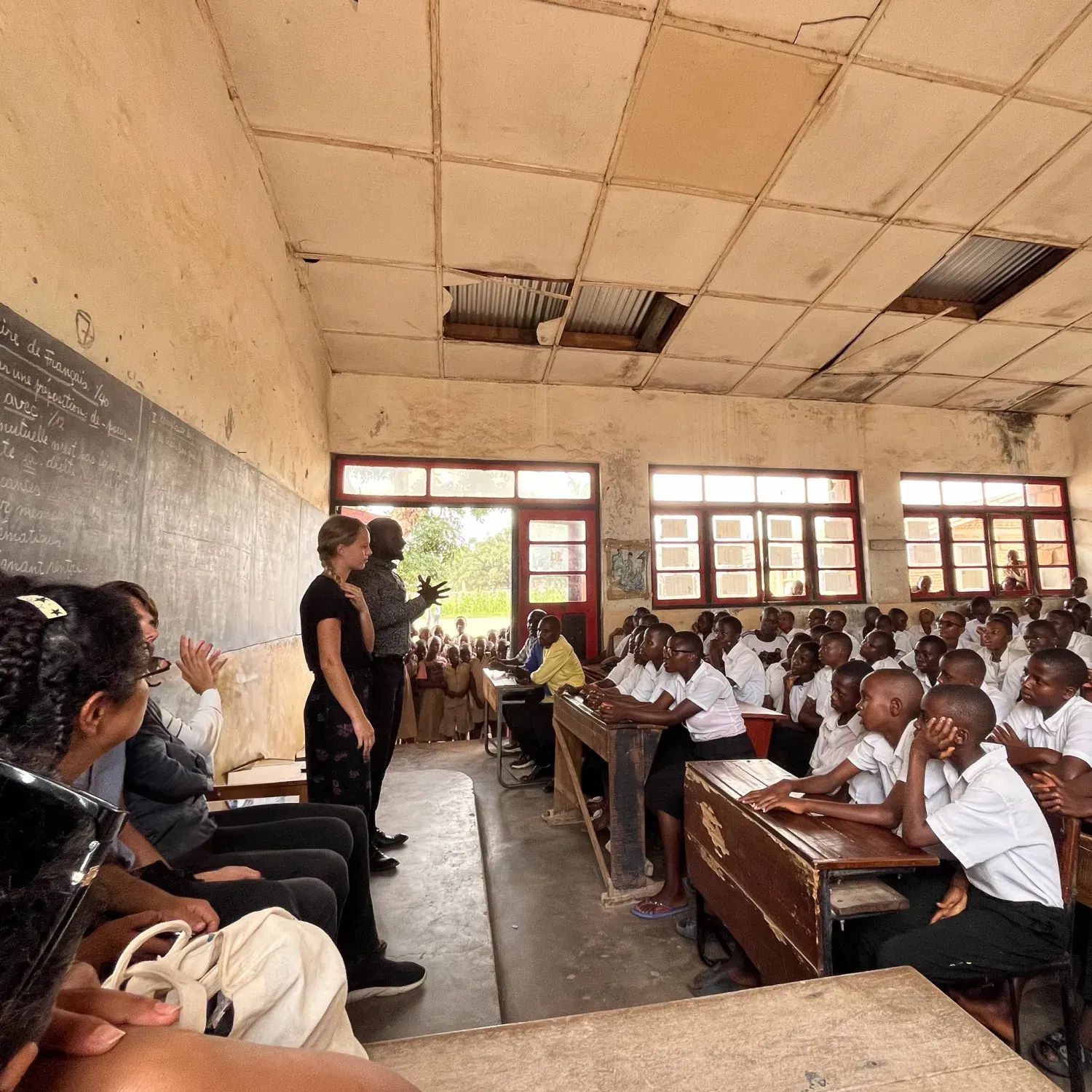 Classroom with many students in white shirts seated at wooden desks attentively listening to two standing adults near a chalkboard in a worn room with broken ceiling tiles.