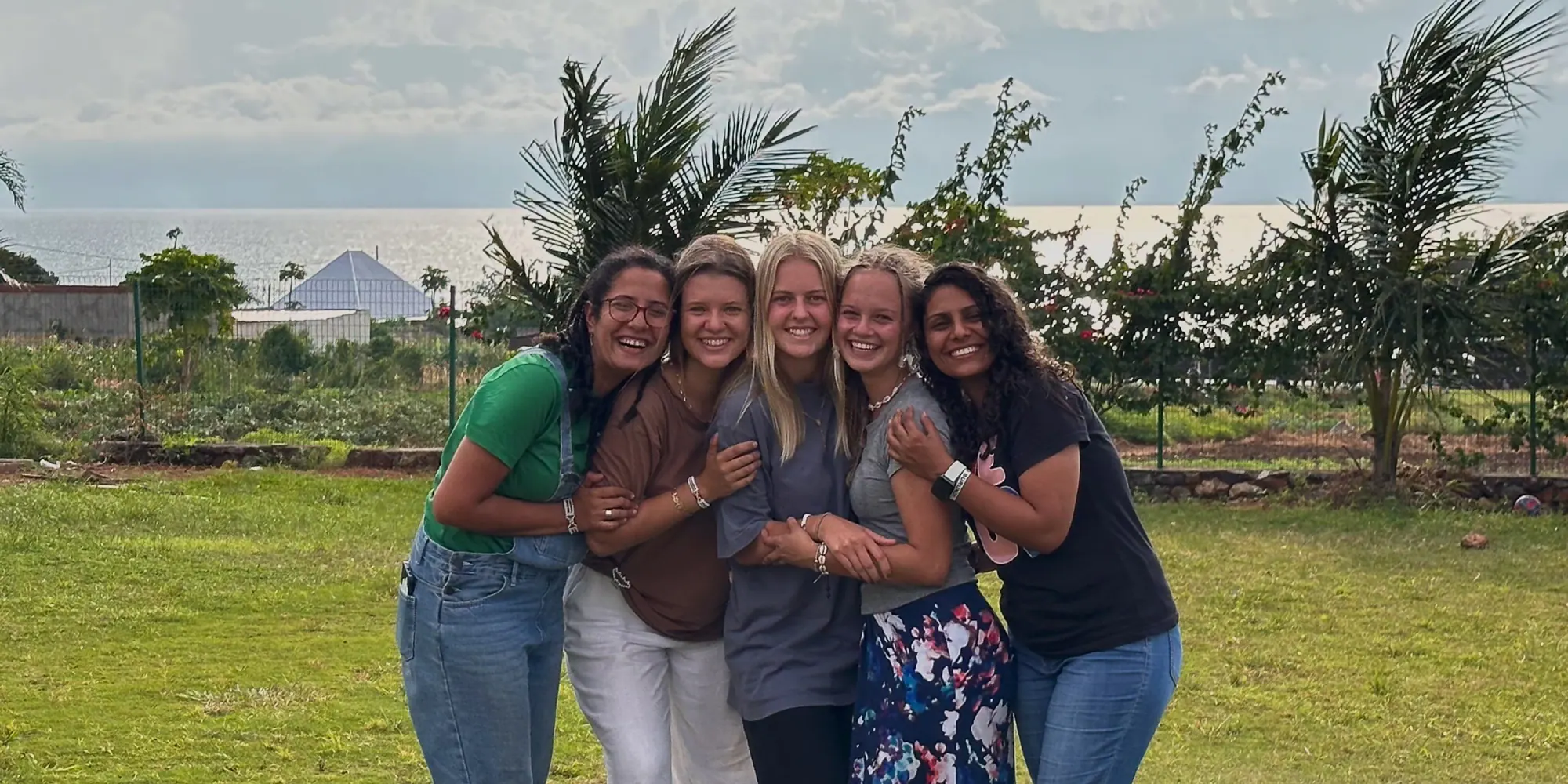 Five young women smiling and embracing each other outdoors on grass with palm trees and the sea in the background.