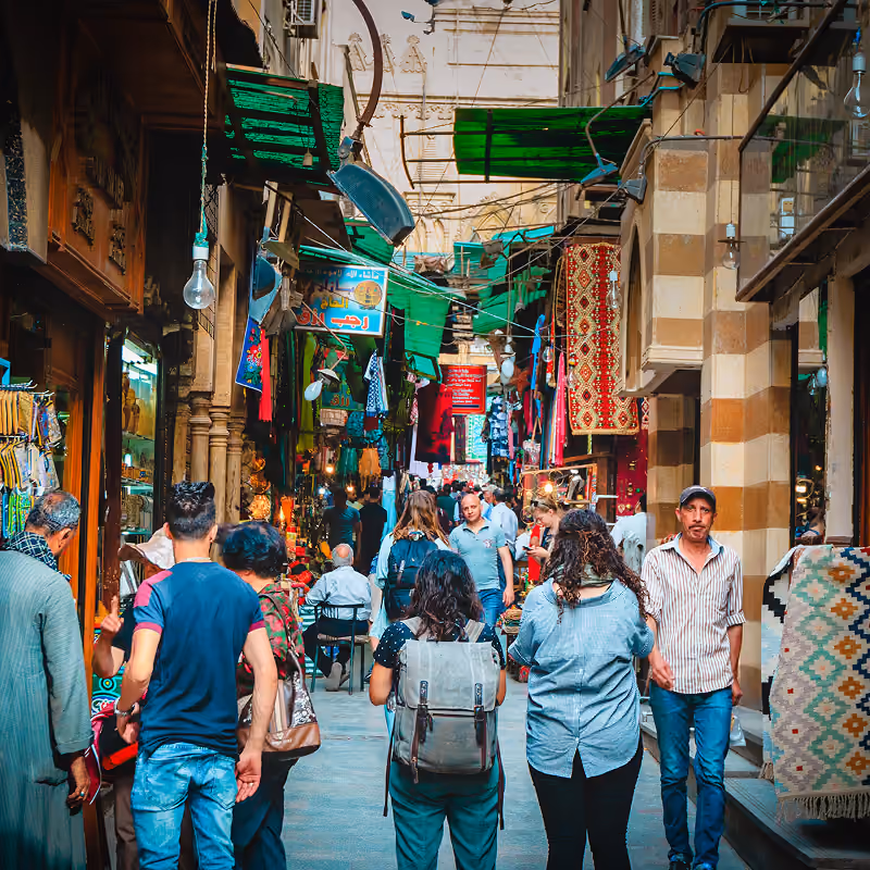 Bustling marketplace street with people walking, colorful fabrics, rugs, and hanging green awnings.