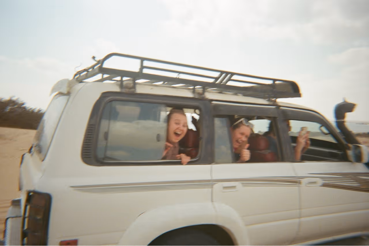 Two people smiling and giving a thumbs up while looking out the windows of a white off-road vehicle.