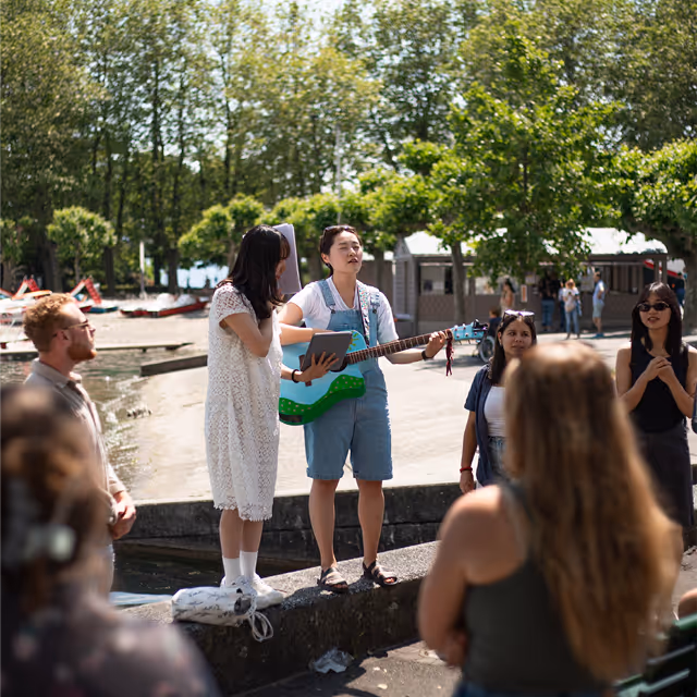 A woman in overalls playing a guitar and singing outdoors while a small group listens, surrounded by trees and sunlight.