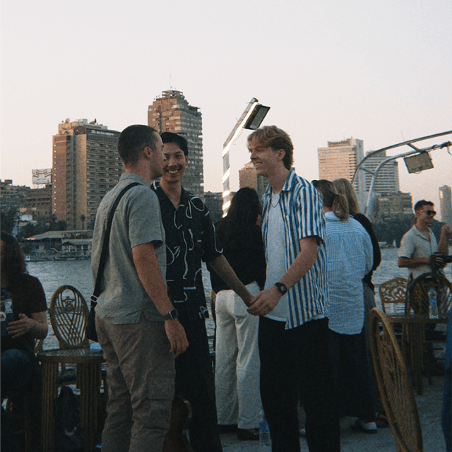 Three young men standing and smiling outdoors near a waterfront with tall buildings in the background during sunset.