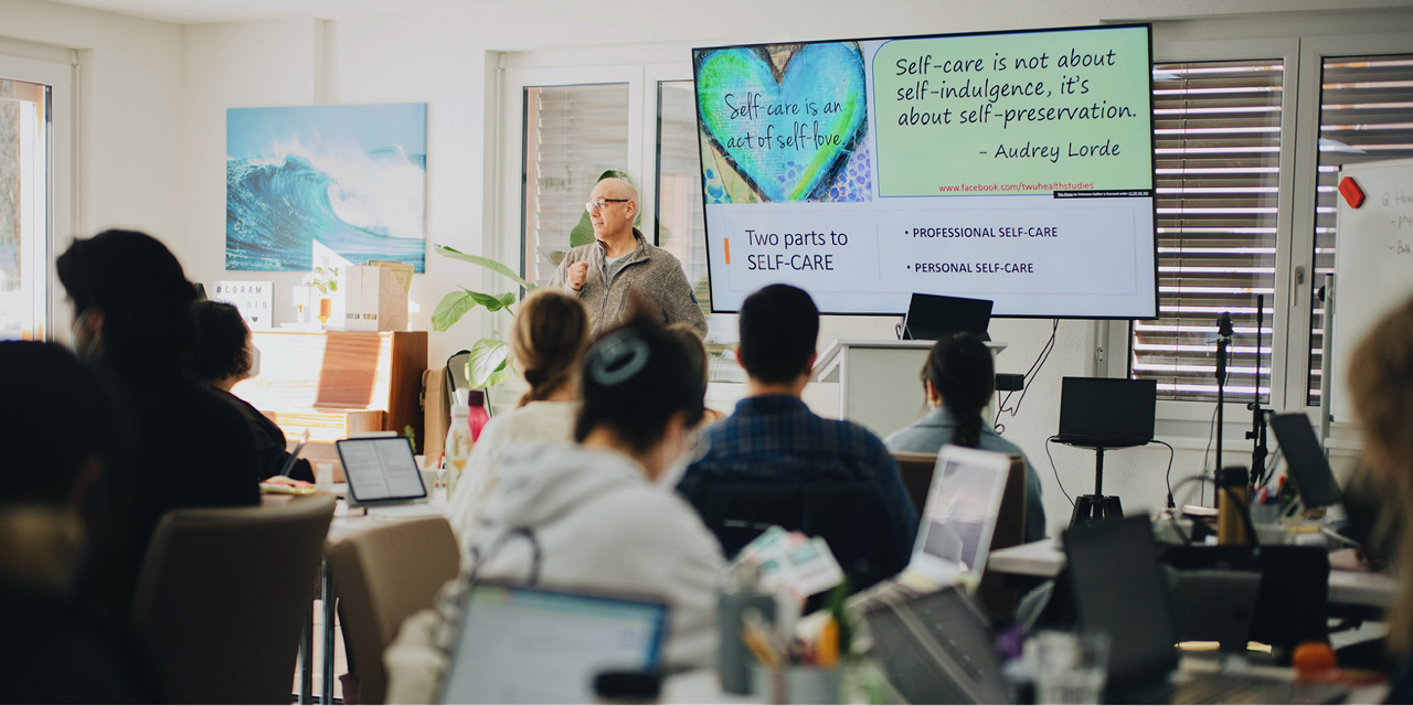Instructor giving a presentation on self-care to an attentive audience in a classroom setting, with a slide showing quotes about self-care and its two parts: professional and personal.