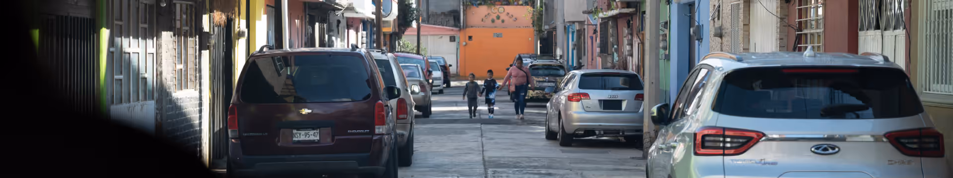 A street with parked cars on both sides and a woman walking hand-in-hand with two children in the distance.