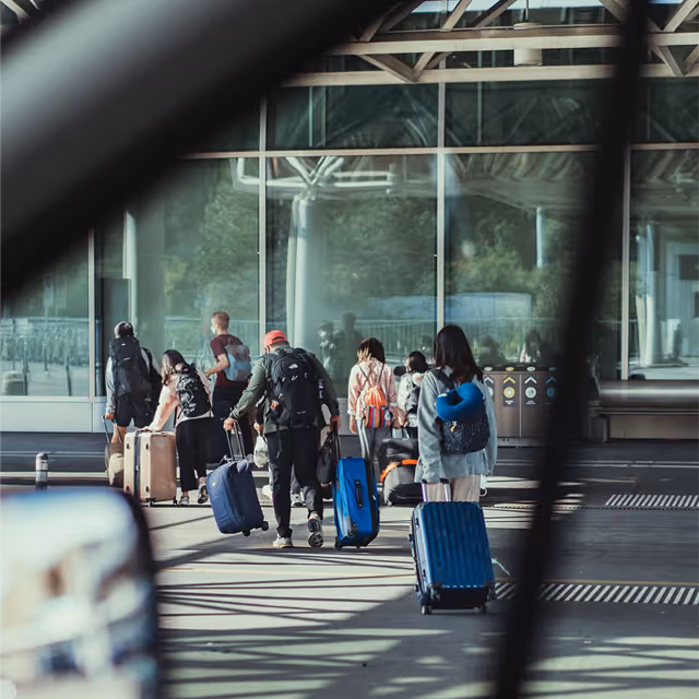 Travelers walking with rolling suitcases near a glass building facade, viewed through a tinted window frame.