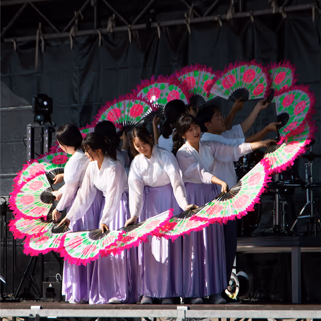 Group of women in white tops and lavender skirts performing a fan dance, holding pink and green fans arranged in a circular pattern on stage.