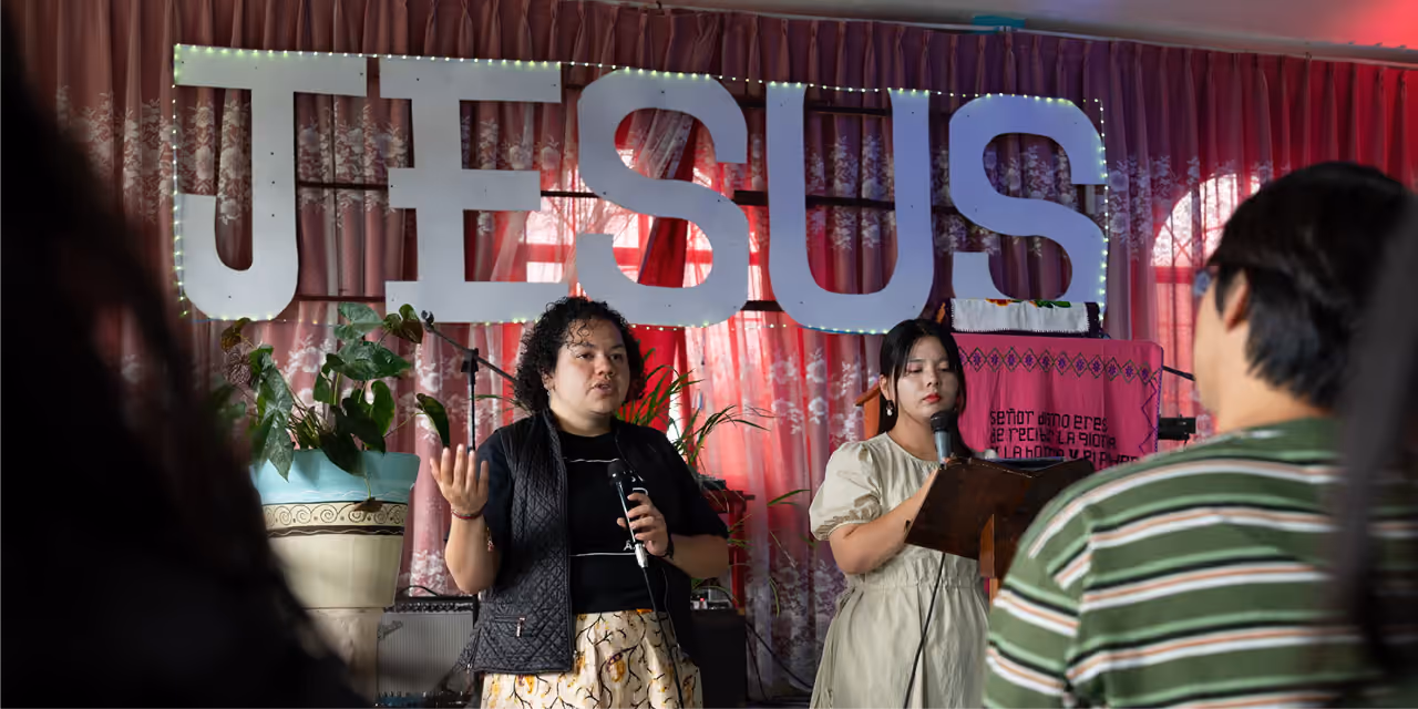 Two women speaking into microphones in front of a large illuminated 'JESUS' sign with an audience in the foreground.
