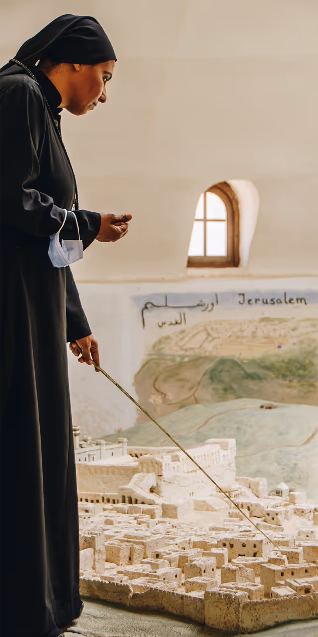 Person dressed in black robe with a head covering pointing at a large stone model of a city labeled Jerusalem.
