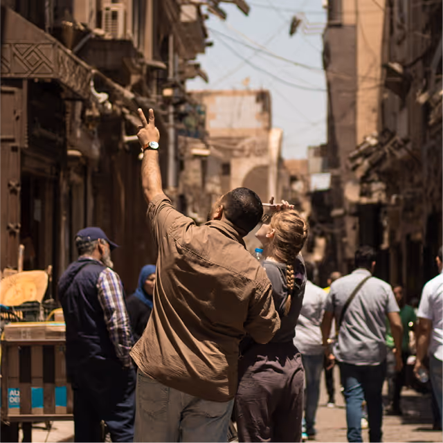Man and woman standing closely on a crowded street with old buildings; man makes a peace sign with his hand raised.