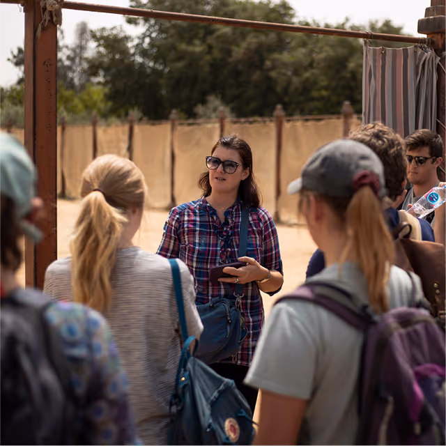 Woman wearing sunglasses and plaid shirt speaking to a group of people outdoors near a sandy area with trees in the background.