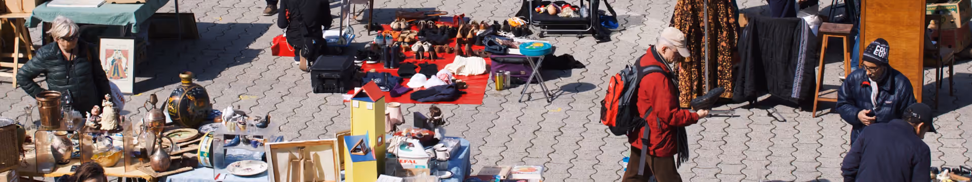 Outdoor flea market scene with tables and blankets displaying various vintage items including ceramics, shoes, and household goods, with several people browsing.