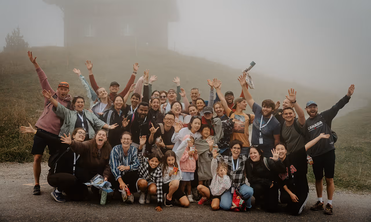 Large diverse group of people outdoors on a foggy day, smiling and raising their hands enthusiastically on a grassy path.