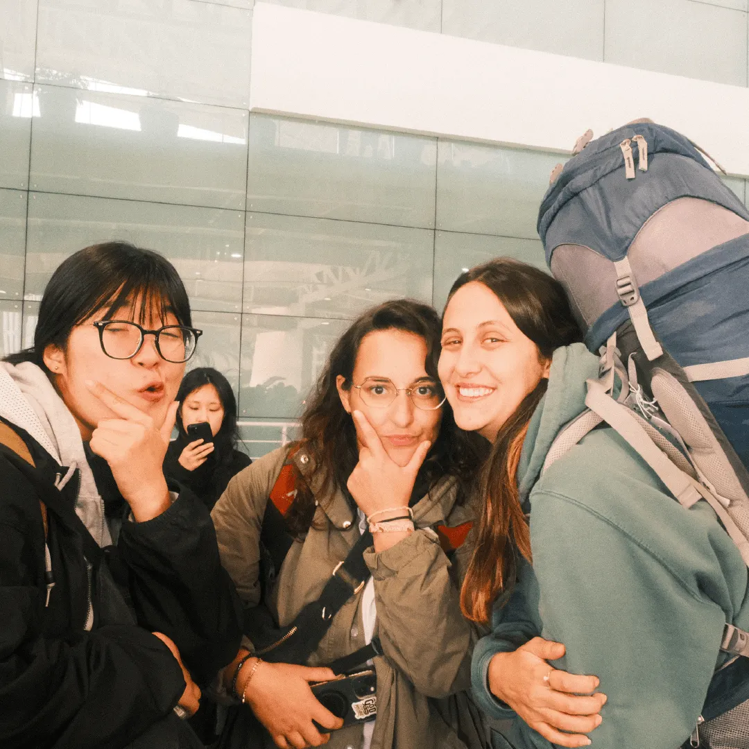Three women posing playfully indoors, one with a large backpack, while a fourth woman in the background looks at her phone.