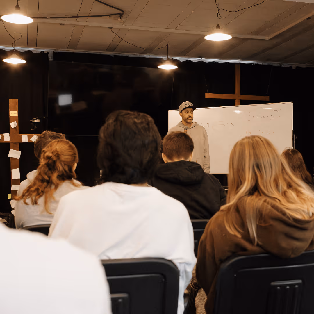 Man wearing a cap and hoodie standing near a whiteboard speaking to a group of seated people in a dimly lit room with crosses on the walls.