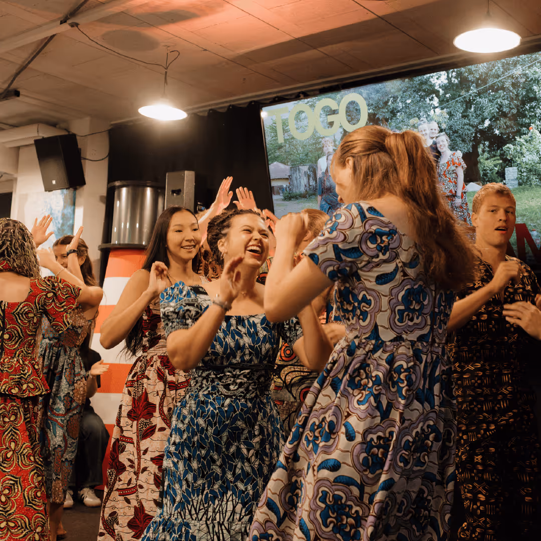 Group of people joyfully dancing indoors wearing colorful patterned dresses and shirts.